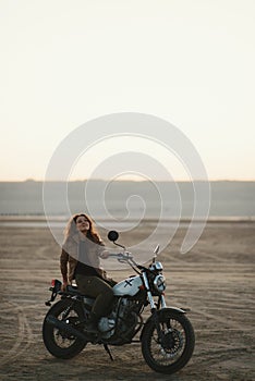 Young beautiful woman sitting on her old cafe racer motorcycle in desert at sunset or sunrise