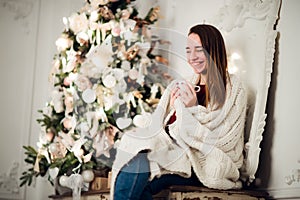 Young beautiful woman with sitting home holding cup of hot coffee wearing knitted warm sweater. Christmas tree