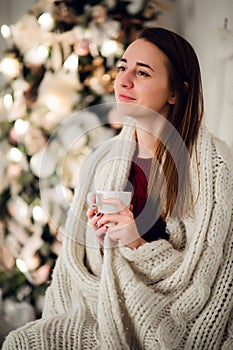 Young beautiful woman with sitting home holding cup of hot coffee wearing knitted warm sweater. Christmas tree