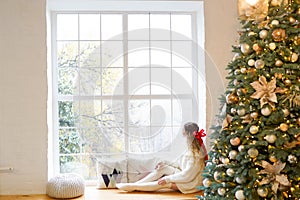 Young beautiful woman sitting home by the window wearing knitted warm sweater. Christmas tree with decorations and lights in the