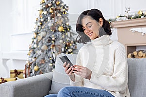 Young beautiful woman sitting near decorated Christmas tree in living room on sofa, smiling and using phone, typing