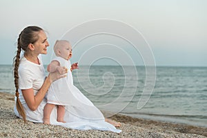 Young beautiful woman sitting on sandy beach and holding baby girl. Mother and child on sea background