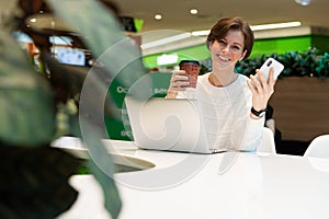 Young beautiful woman sitting at a shopping center at a table and working at a computer laptop, using mobile phone