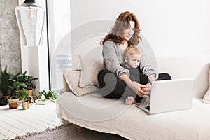 Young beautiful woman sitting on sofa with her little handsome son dreamily using laptop together. Mom with baby boy