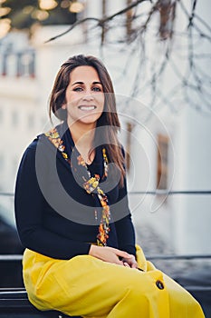 Young beautiful woman is sitting on the street on the bench at sunset. Portrait of a smiling girl in a yellow skirt, modern sneake