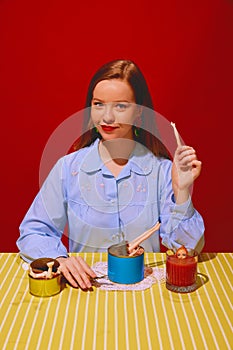 Young beautiful woman sitting at table and eating canned meat, drinking tomato juice against red background. Quirky food