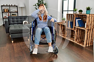 Young beautiful woman sitting on wheelchair at home smiling and laughing with hand on face covering eyes for surprise