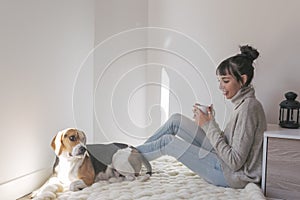 young beautiful woman sitting on a white blanket and holding a cup of coffee. cute beagle dog besides. Relaxing at home. Indoors