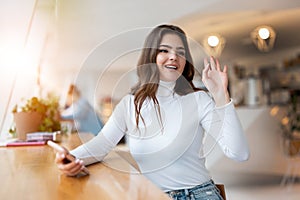 Young beautiful woman with smarphone in her hand waving her friend l while sitting in the cafe during lunch break, positive vibes photo