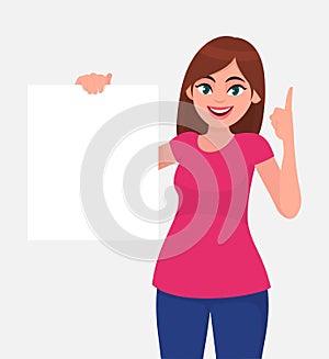 Young beautiful woman smiling and holding a blank / empty sheet of white paper or board and pointing up finger.