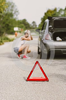 Young beautiful woman spection a rear tire on the side of the road. Car with problems and red triangle to warn other road users