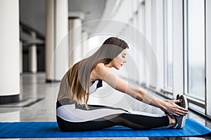 Young beautiful woman in sportswear doing stretching while sitting on the floor in front of window at gym