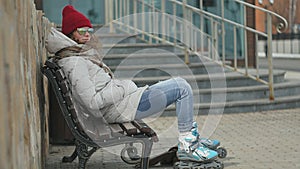 Young beautiful woman in sporty warm clothes and rollers, sitting on a wooden bench and dresses roller skates getting