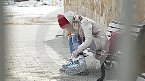 Young beautiful woman in sporty warm clothes and rollers, sitting on a wooden bench and dresses roller skates getting