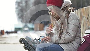 Young beautiful woman in sporty warm clothes and rollers, sitting on a wooden bench and dresses roller skates getting