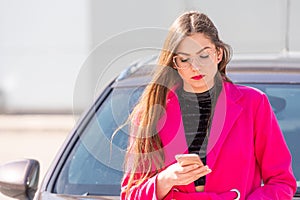 A young, beautiful woman standing by the car and writing a text message on her smartphone