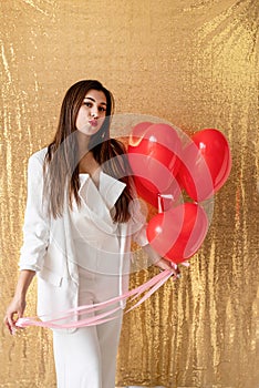Young beautiful woman standing on golden shiny background holding red heart chaped balloons