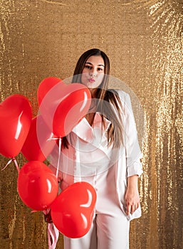 Young beautiful woman standing on golden shiny background holding red heart chaped balloons