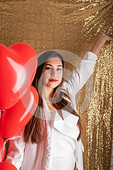 Young beautiful woman standing on golden shiny background holding red heart chaped balloons