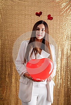 Young beautiful woman standing on golden shiny background holding red heart chaped balloons