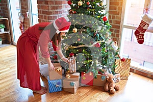 Young beautiful woman standing with happy face picking up present from under the christmas tree wearing santa claus hat