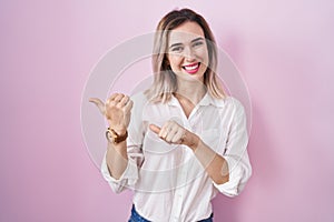 Young beautiful woman standing over pink background pointing to the back behind with hand and thumbs up, smiling confident