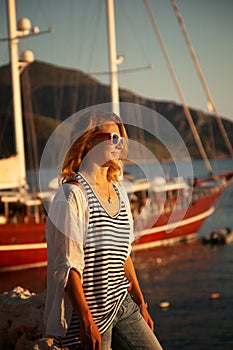Young beautiful woman standing on the seashore next to a yacht at sunset, vacation and summer sea travel