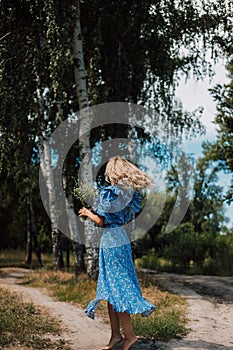 A young beautiful woman stands with her back in the woods in a long blue dress