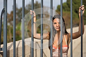 Young and beautiful woman with straight brown hair, orange top, clinging to the bars of a fence, with a sensual and flirtatious