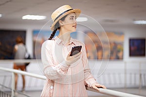 Young beautiful woman in straw hat is holding smartphone. In background is art exhibition hall of gallery with paintings