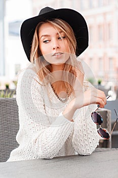 Young beautiful woman in a stylish white knitted sweater in an elegant black hat with sunglasses is sitting at a table