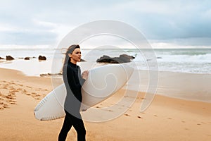 Young beautiful woman with surfboard walking on beach at sunny day, enjoying seaside and active recreation, copy space