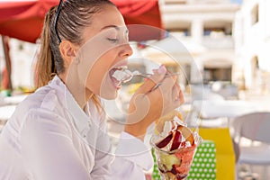 Young beautiful woman takes a break in her shopping day in city eating a big ice cream sitting at cafe bar dehore. Vices and