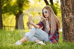 Young beautiful woman takes selfie on cell phone sitting on grass in summer city park. Blow kiss. Beautiful modern girl in