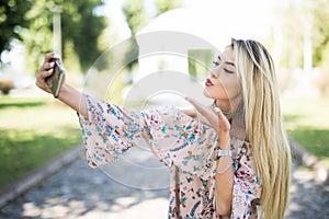 Young beautiful woman takes selfie on cell phone sitting on grass in summer city park. Blow kiss. Outdoor