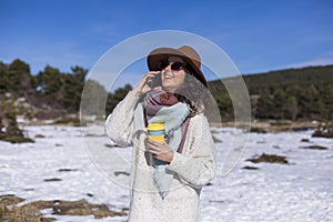 Young beautiful woman talking on her mobile phone in the snow and holding a cup of tea or coffee. Outdoors portrait, lifestyle