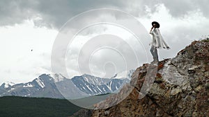 Young beautiful woman traveler wearing hat and poncho relaxing on the top of the hill with mountains and hills around.