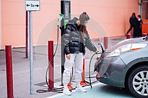 Young beautiful woman traveling by electric car having stop at charging station