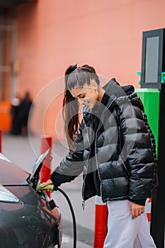 Young beautiful woman traveling by electric car having stop at charging station