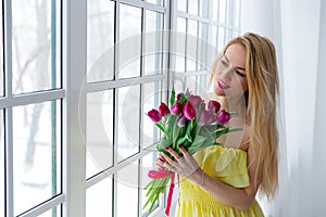 Young beautiful woman with tulip bunch in yellow dress looks at window.