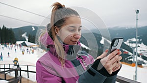 Young beautiful woman uses a mobile phone on the lift of a ski resort, mountains and skiers in the background.