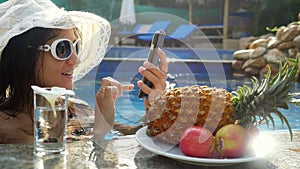 Young beautiful woman uses mobile phone in swimming pool and drinks water next to plate with tropical fruits. 3840x2160