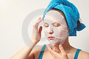A young beautiful woman uses a moisturizing cosmetic fabric face mask with a towel wrapped around her head. on white background