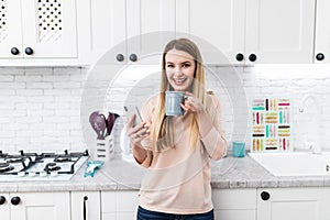 Young beautiful woman using cell phone and having a coffee in the kitchen
