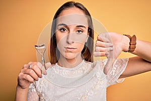 Young beautiful woman using eyelash curler standing over isolated yellow background with angry face, negative sign showing dislike