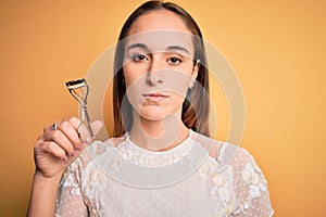 Young beautiful woman using eyelash curler standing over isolated yellow background with a confident expression on smart face