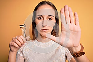 Young beautiful woman using eyelash curler standing over isolated yellow background with open hand doing stop sign with serious