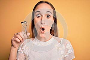 Young beautiful woman using eyelash curler standing over isolated yellow background scared in shock with a surprise face, afraid