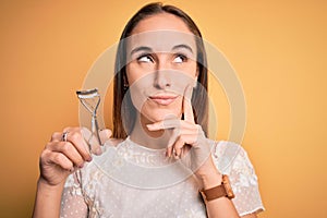 Young beautiful woman using eyelash curler standing over isolated yellow background serious face thinking about question, very