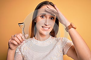 Young beautiful woman using eyelash curler standing over isolated yellow background stressed with hand on head, shocked with shame